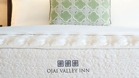 A white quilted mattress with the text 'OJAI VALLEY INN' printed on the side, styled on a bed with white pillows and a green patterned accent pillow in front of a beige upholstered headboard.