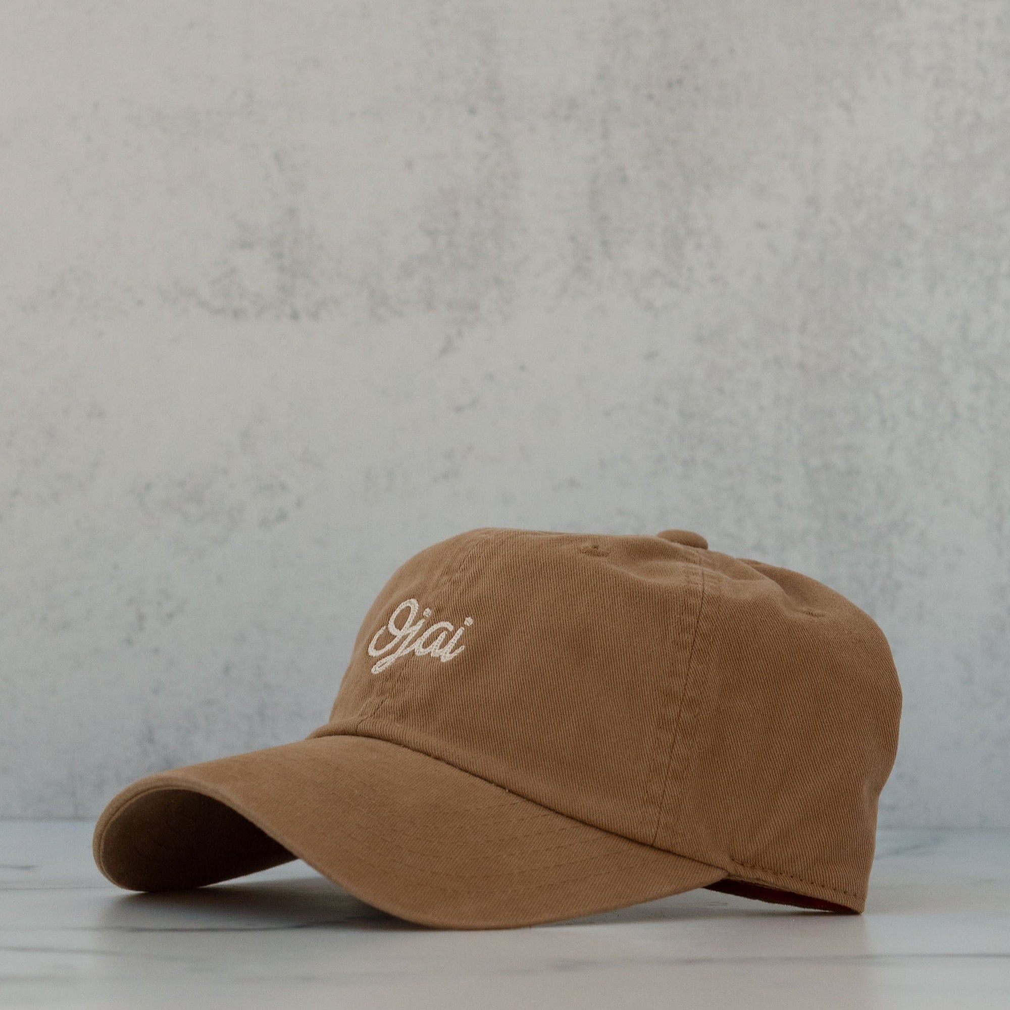 A khaki-colored, slouch-style hat with a curved brim and a self-fabric strap, displayed against a textured background.