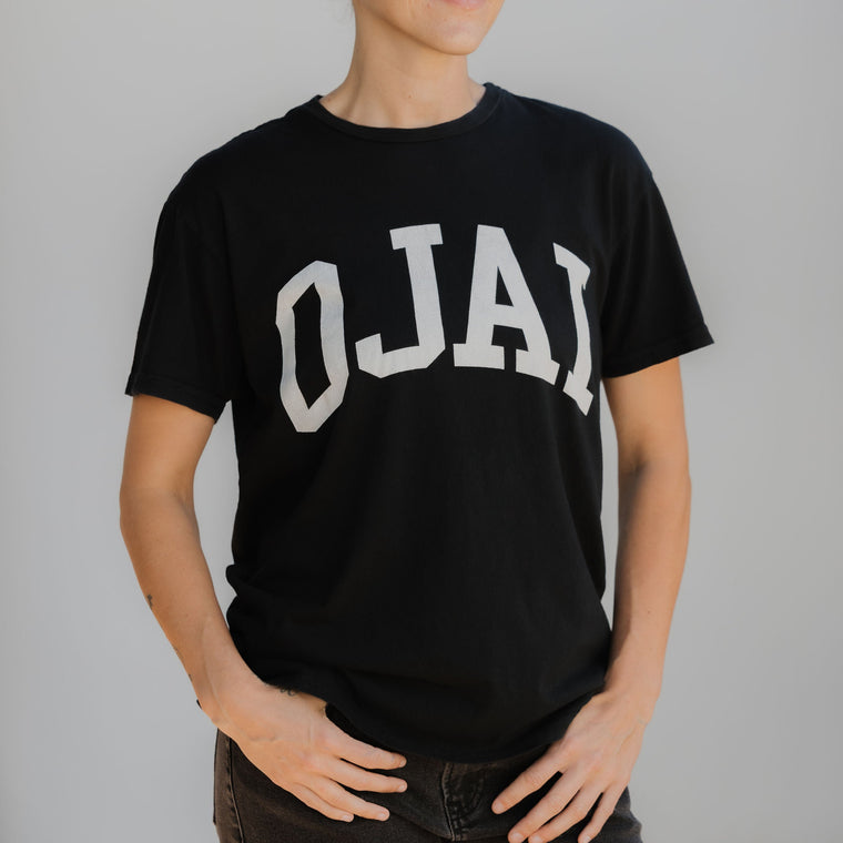 Person wearing a black t-shirt with 'OJAI' printed on it against a plain background