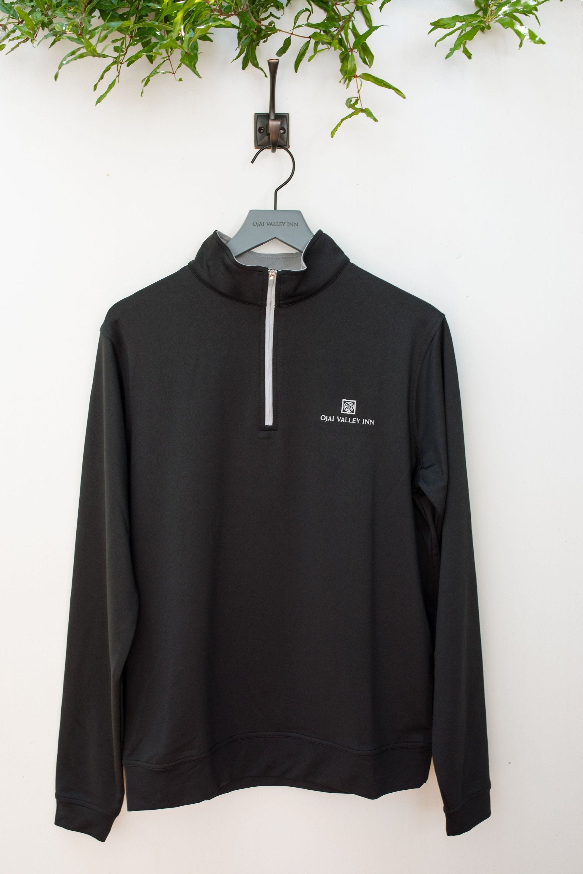 A black Peter Millar Perth Performance 1/4 zip jacket hanging on a hook, with a mock collar and banded cuffs and hem.