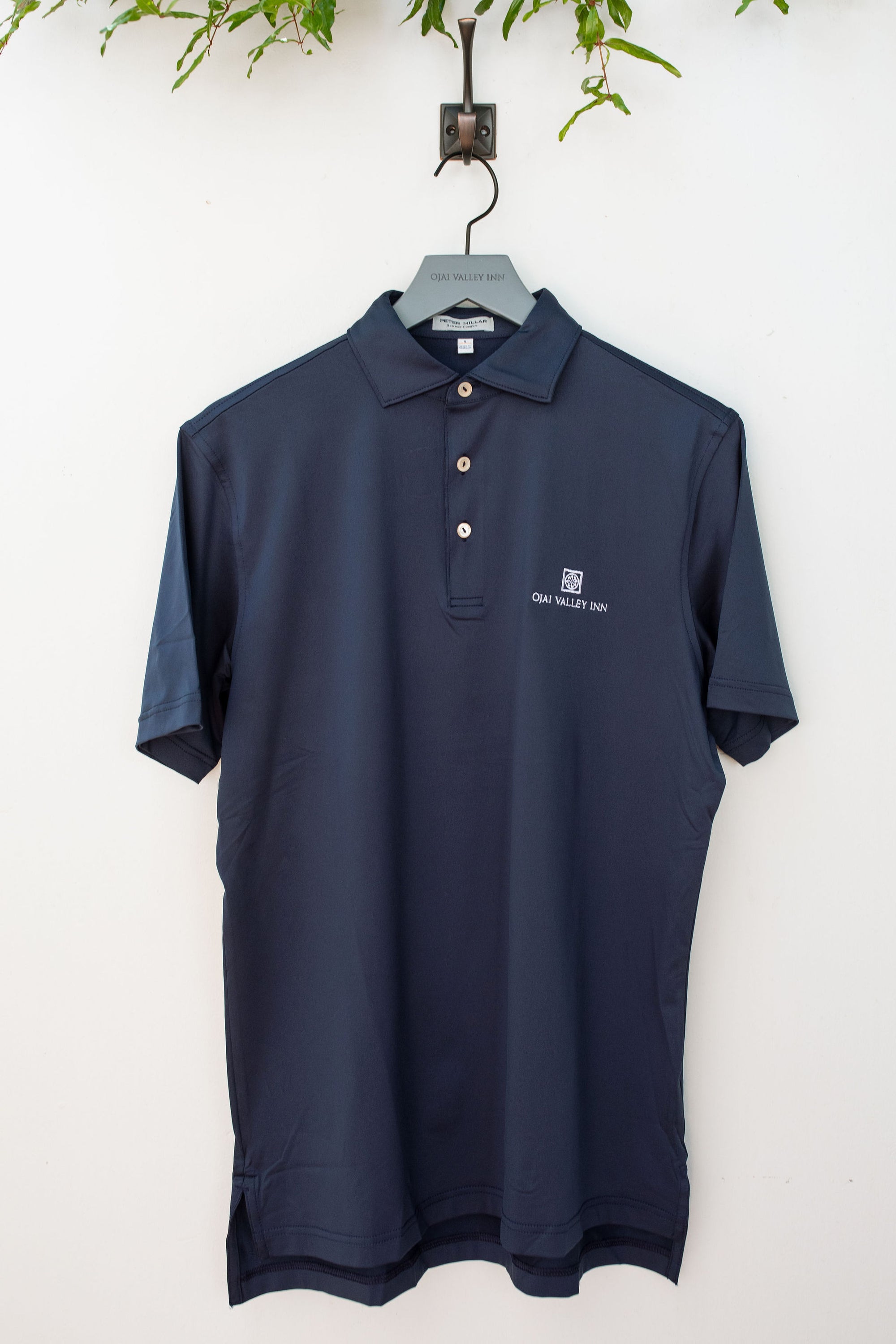 A navy-colored Peter Millar Solid Performance Jersey Polo shirt hung on a hanger against a white wall, with a three-button placket and a collar visible.