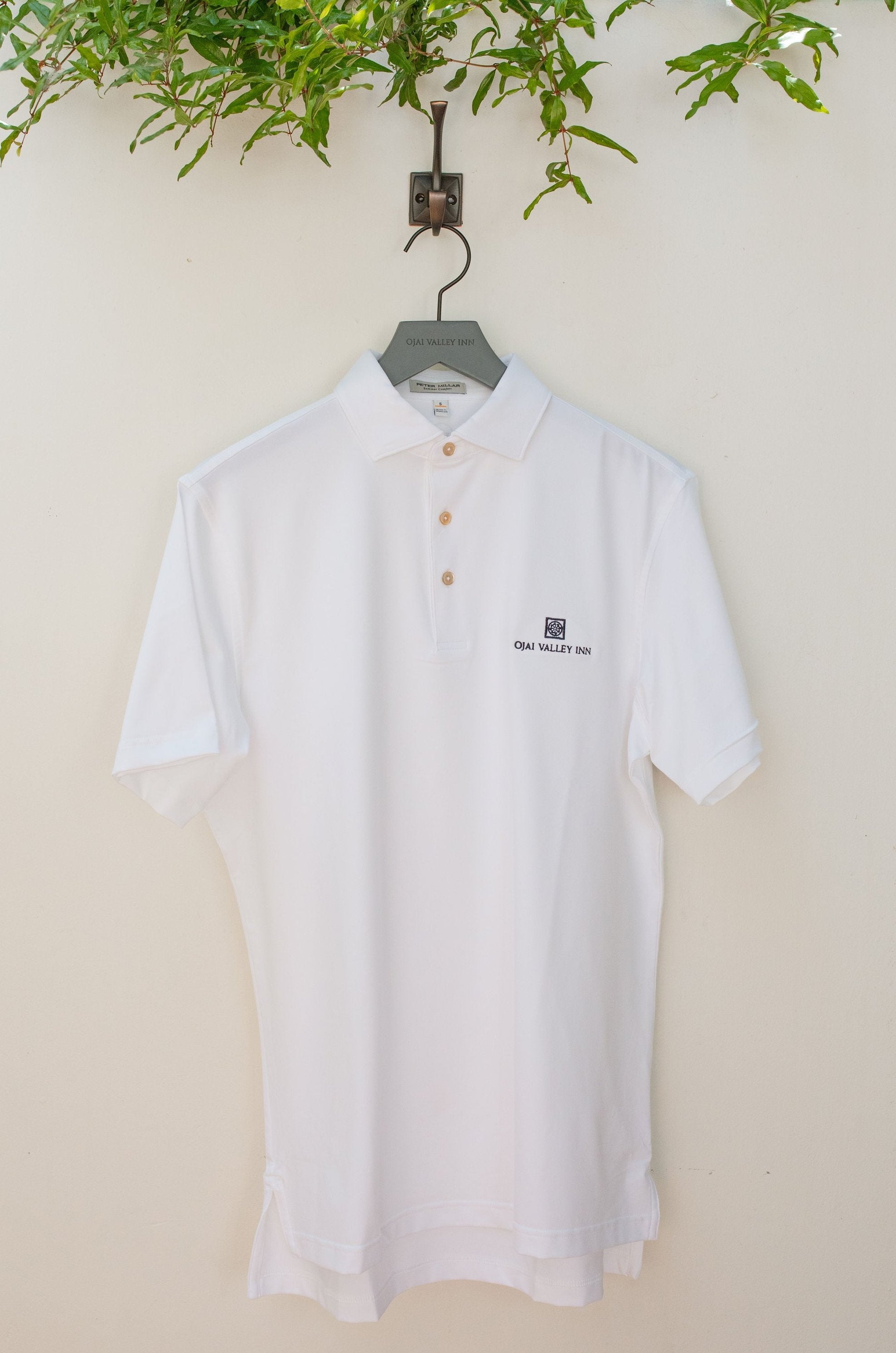 A white, short-sleeved Peter Millar polo shirt hung on a hanger against a wall, with a green potted plant and a grey hook above it.