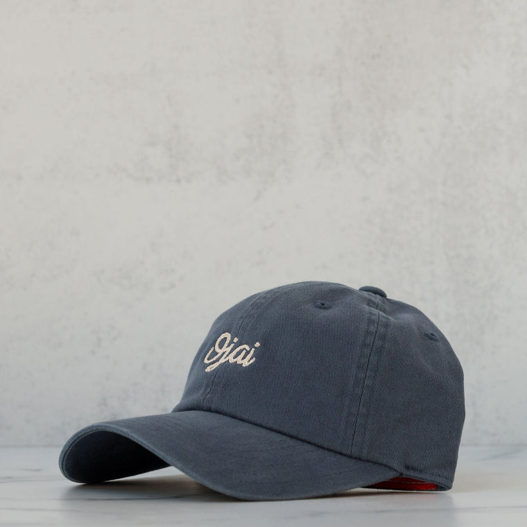 A cloud blue, slouch-style hat with a curved brim and a self-fabric strap, displayed against a textured grey background.