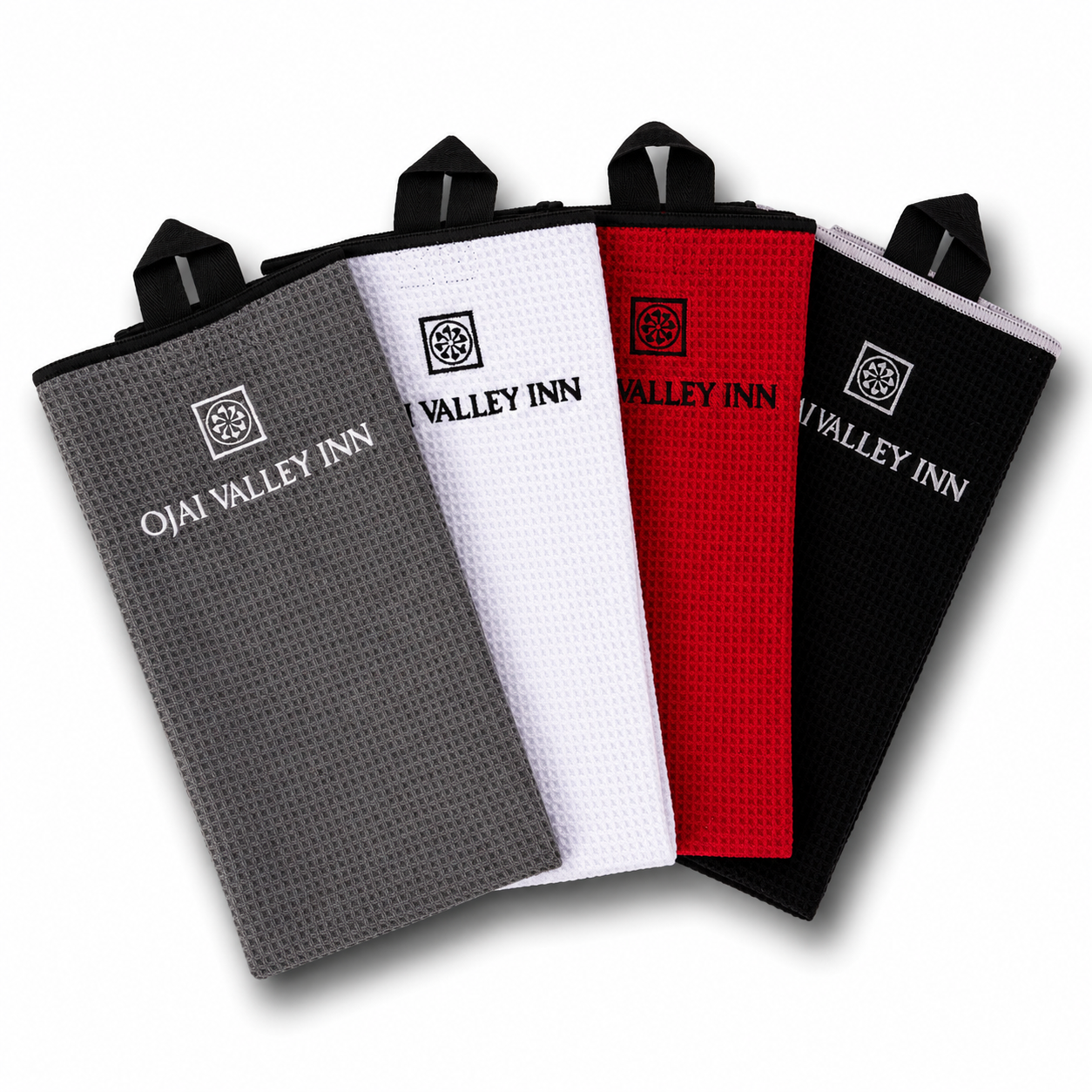 Four textured golf towels in gray, white, red, and black, arranged in a fan shape. Each towel features the 'Ojai Valley Inn' logo and name printed in white, along with a black hanging loop at the top.