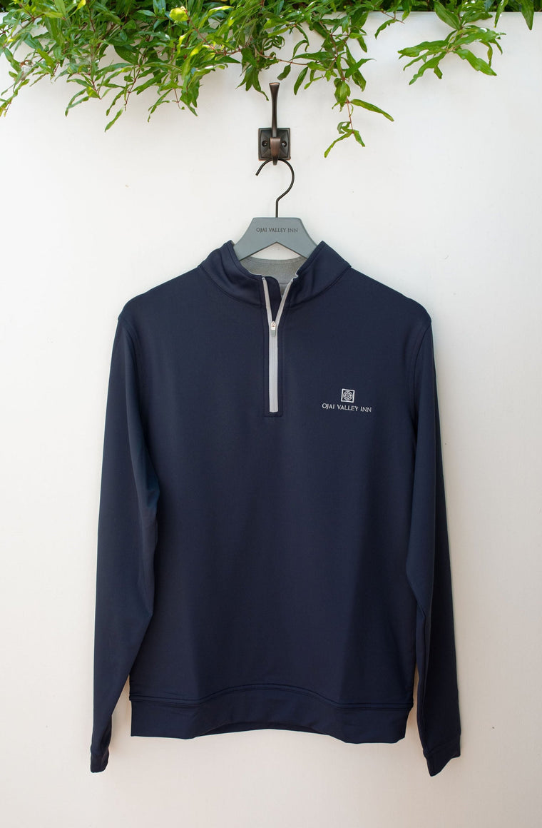 A navy-colored, quarter-zip performance golf jacket hanging on a hook, with a mock collar and banded cuffs, displayed against a white wall with green foliage in the background.