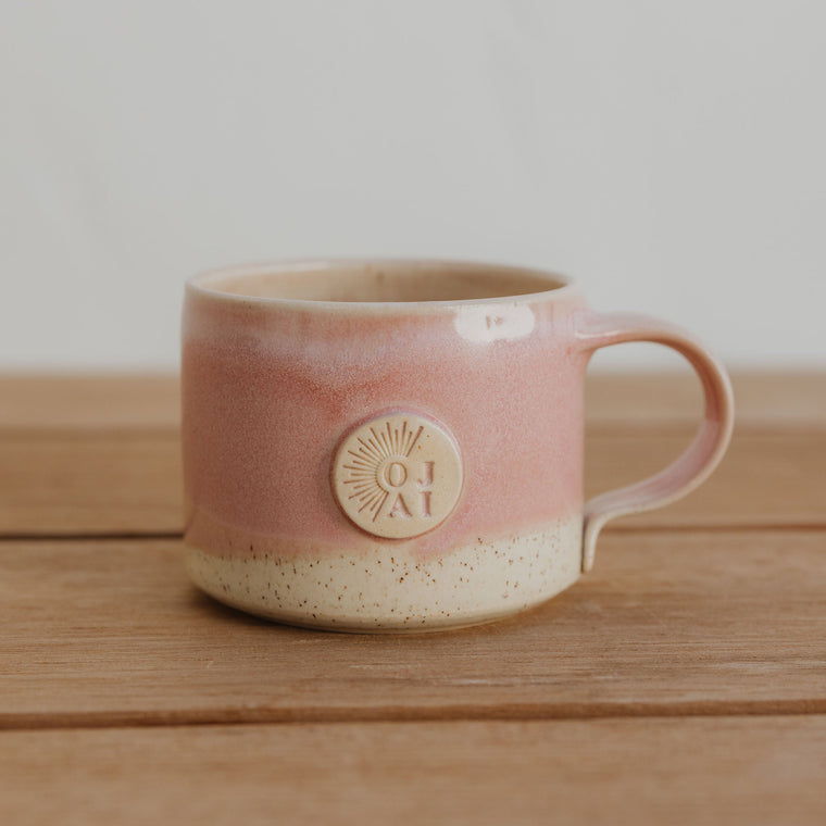 Pink ceramic mug with a circular logo that says 'Ojai' on the front, on a wooden surface