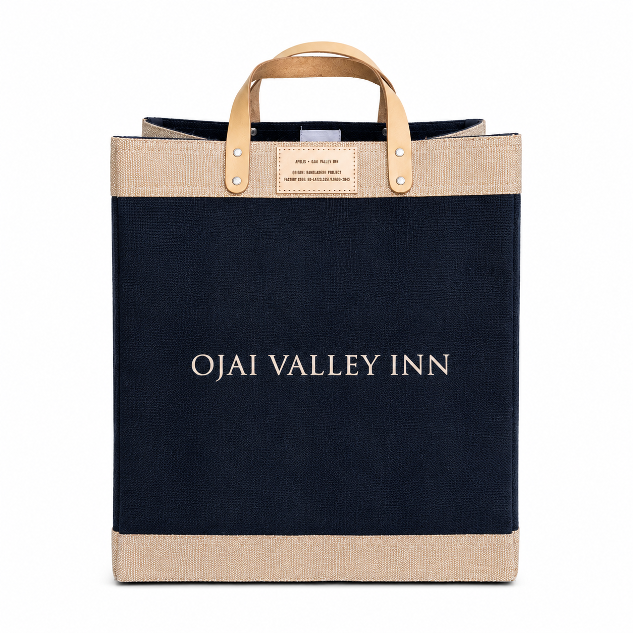 A canvas tote bag from Ojai Valley Inn featuring a dark navy blue body, natural beige jute trim at the top and bottom, and light tan leather handles with metal rivets. The words 'OJAI VALLEY INN' are printed in white serif font across the center.