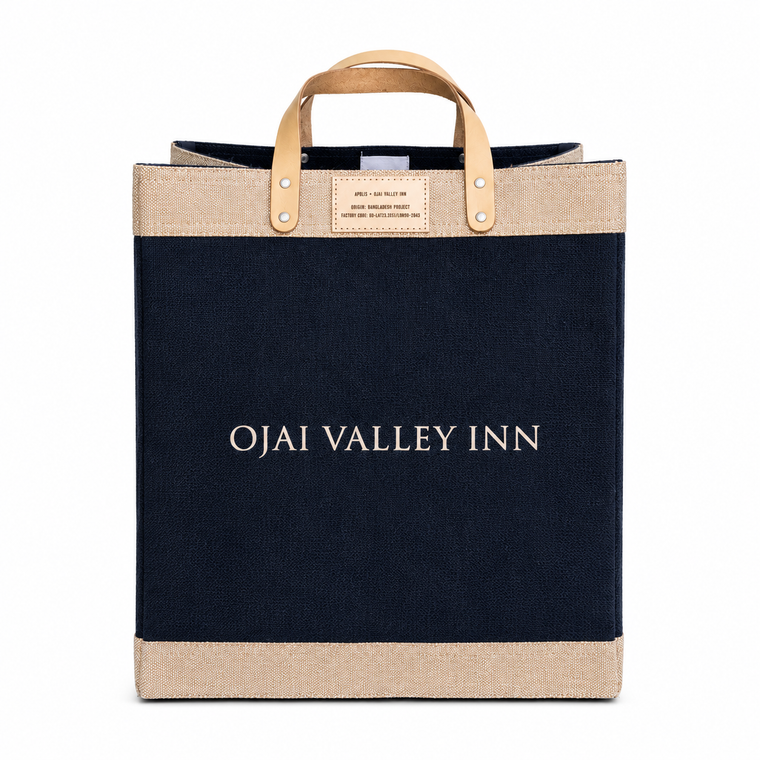 A canvas tote bag from Ojai Valley Inn featuring a dark navy blue body, natural beige jute trim at the top and bottom, and light tan leather handles with metal rivets. The words 'OJAI VALLEY INN' are printed in white serif font across the center.