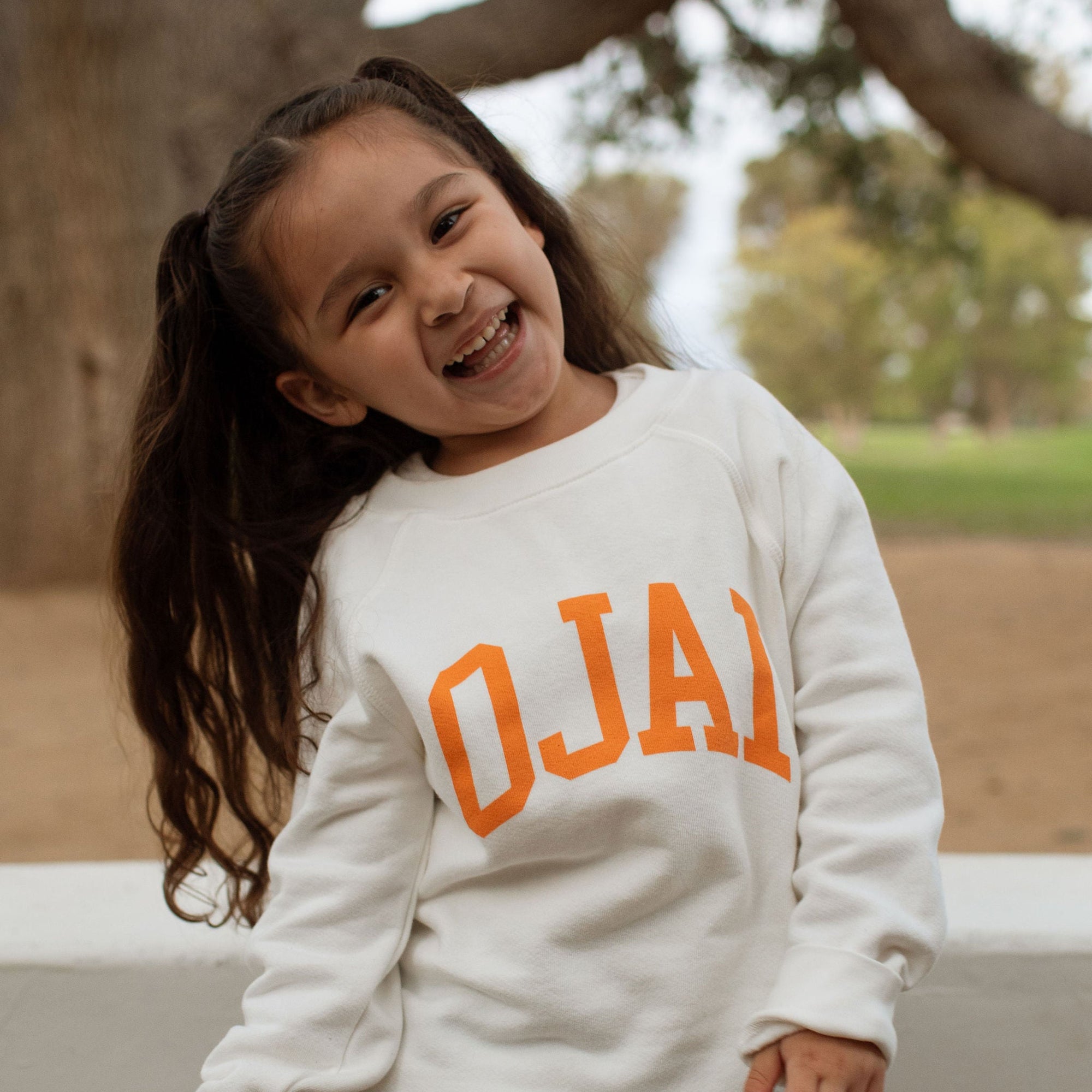 A young girl with long dark hair in a ponytail is smiling and looking to the side, wearing a cream-colored crewneck sweatshirt with 'OJAJ' printed in orange across the chest, paired with light blue jeans, set against a blurred outdoor background with trees.
