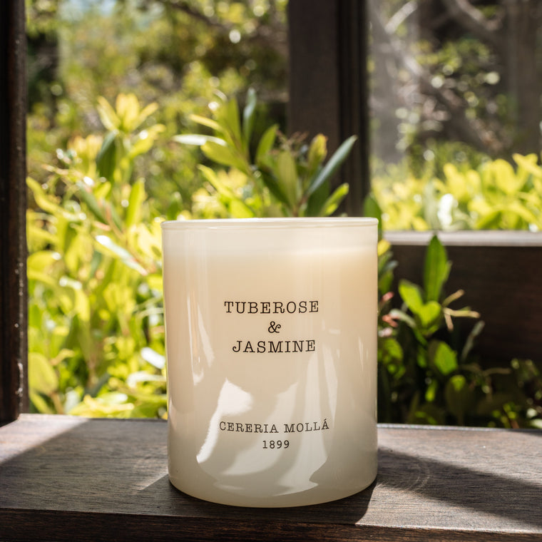 A frosted glass candle jar labeled 'TUBEROSE & JASMINE' and 'CINERARIA MOLLIS 1899' sits on a dark wooden windowsill with a blurred garden background.