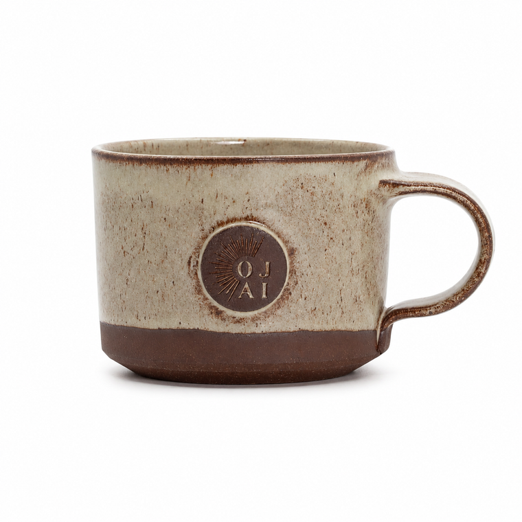 A handmade ceramic mug with a speckled beige and brown glaze, featuring a circular embossed logo on the front that reads 'OJ AI'.