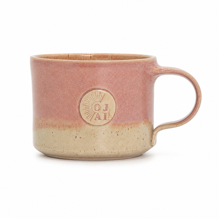 A handmade ceramic mug with a dusty rose pink upper body and a textured beige lower body, featuring a circular embossed logo on the front.