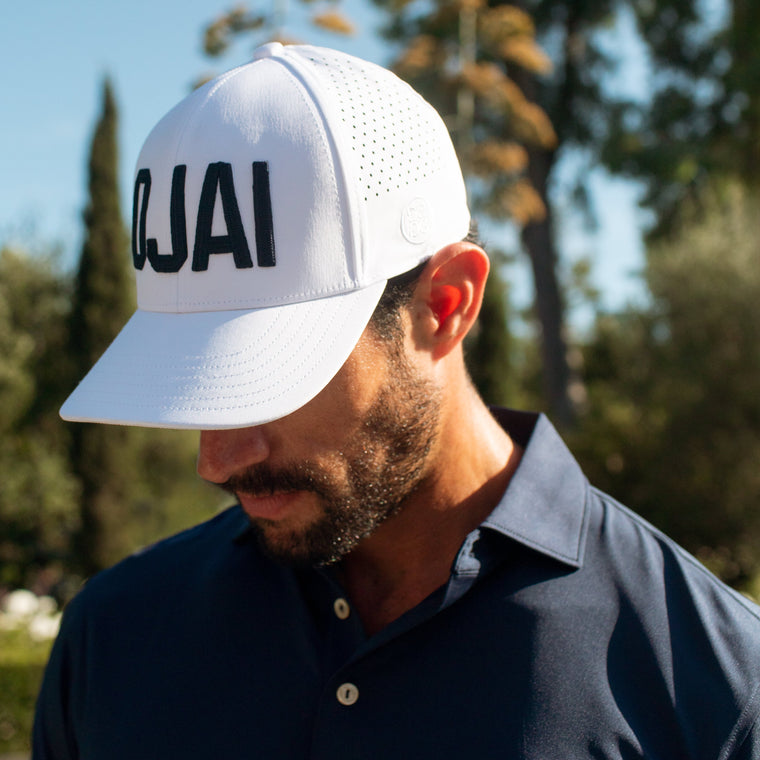 A man wearing a white Ojai trucker cap with black lettering and a navy blue polo shirt with the Ojai Valley Inn logo, photographed outdoors with trees and blue sky in the background.