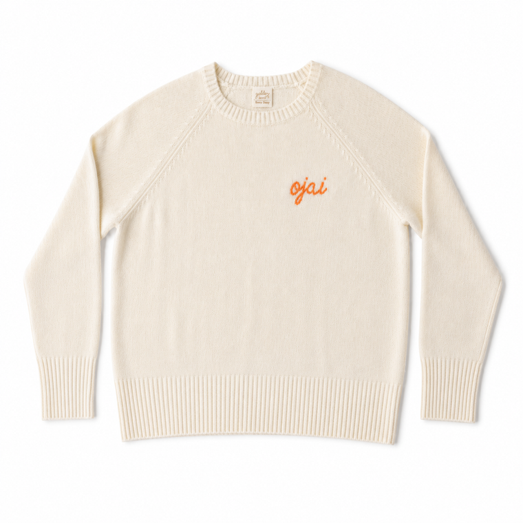 A cream-colored knit crewneck sweater with 'ojai' embroidered in orange on the left chest, displayed flat against a white background.