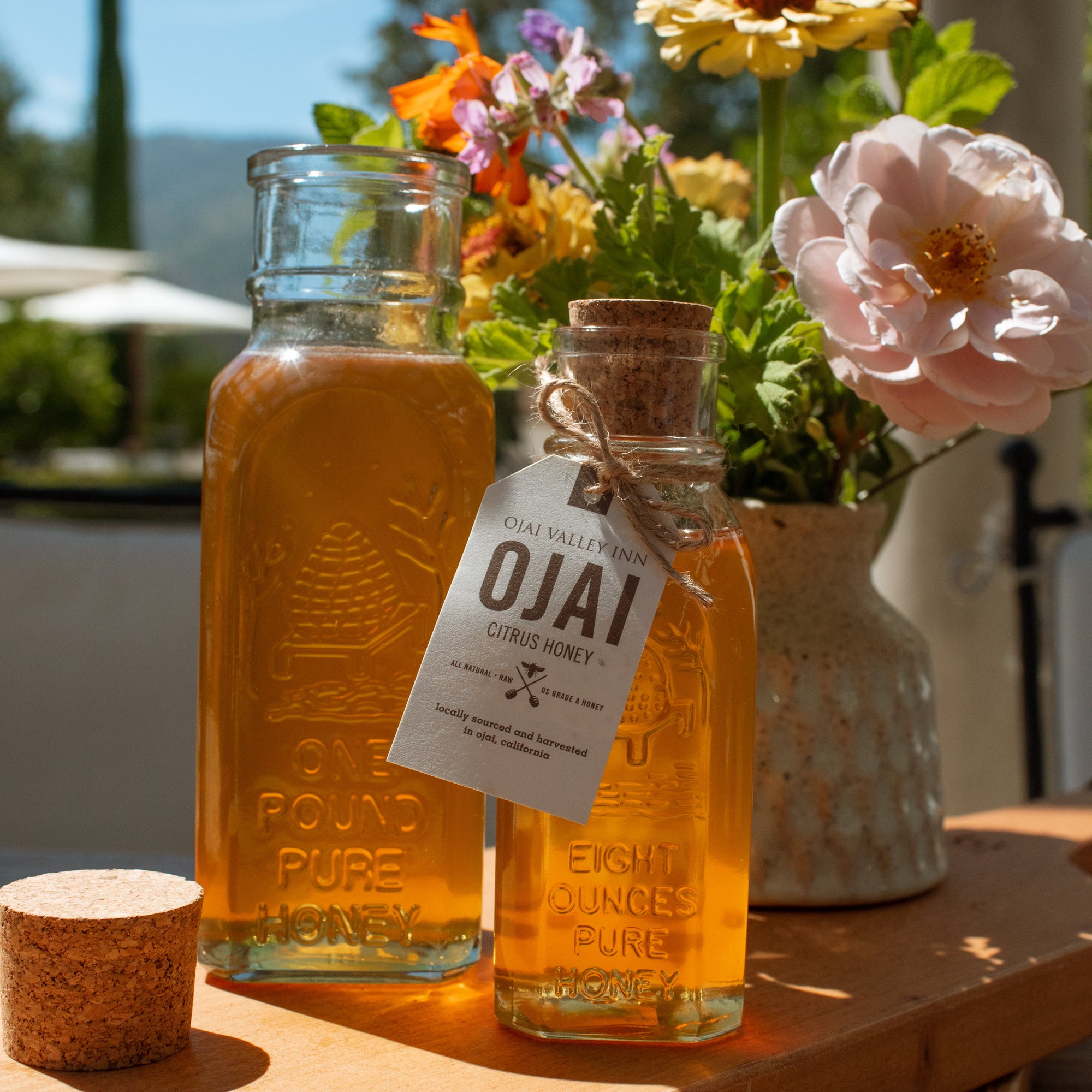 Two glass bottles of pure honey on a wooden tray outdoors, with a vase of colorful flowers behind them. One bottle has a cork stopper and a hanging tag that reads 'OJAI'.