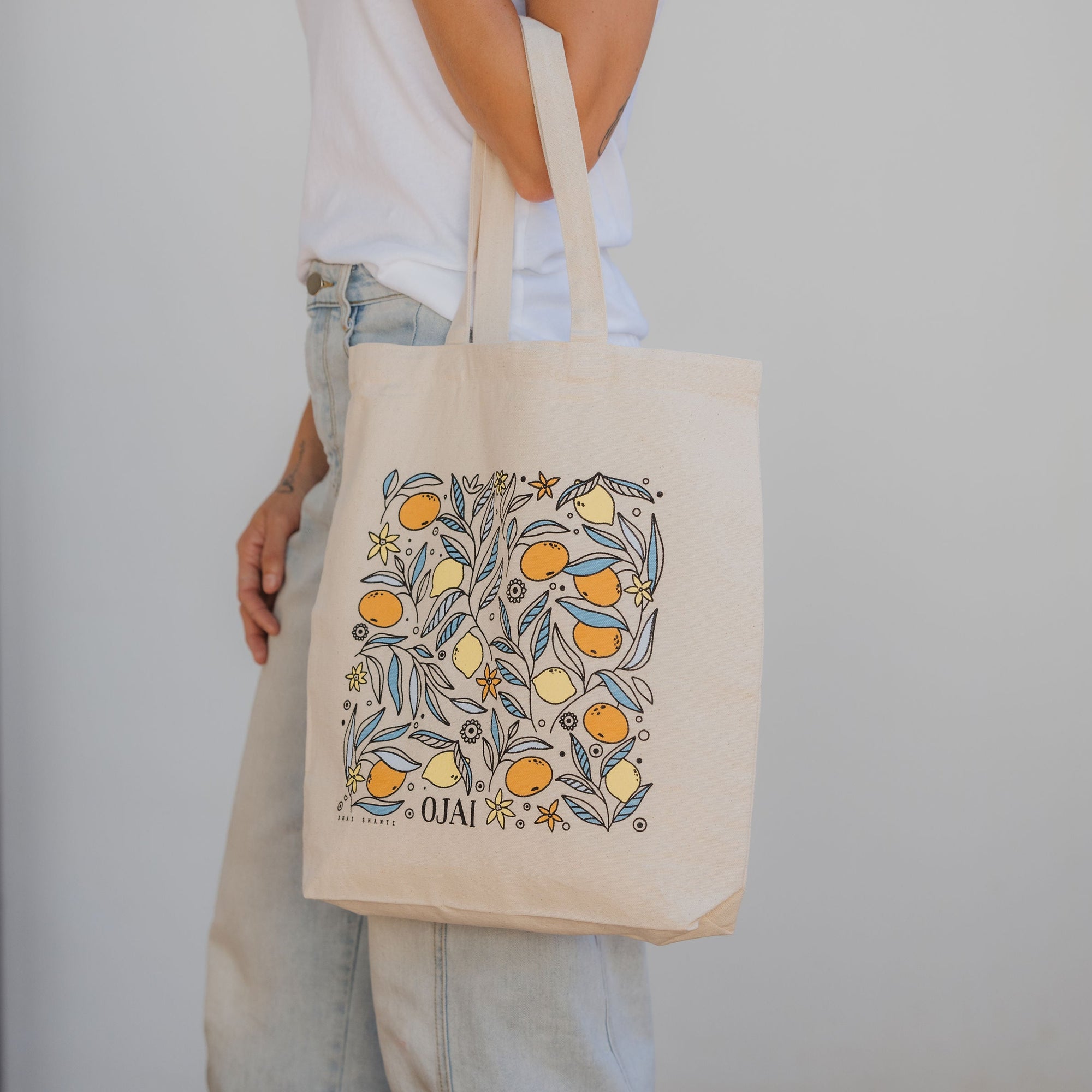 Person holding a tote bag with a floral design and 'Ola!' text against a plain background