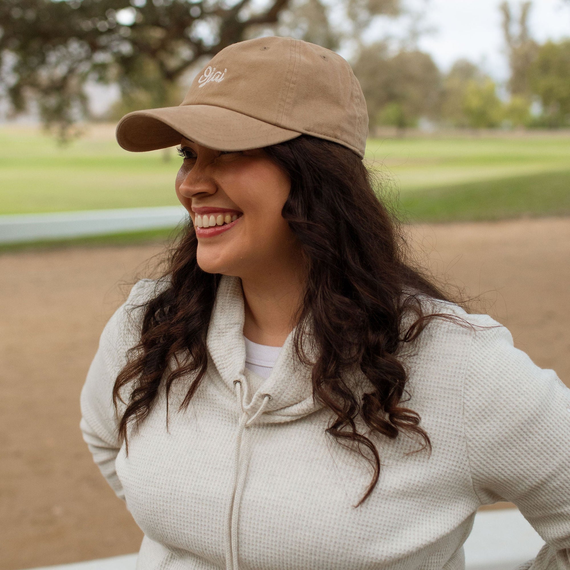 A woman smiling outdoors on a golf course, wearing a tan baseball cap with a small embroidered logo and a light beige zip-up hoodie.