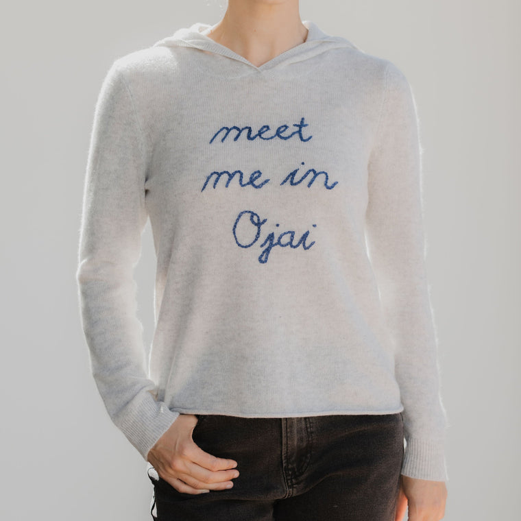 person wearing grey hoodie that reads 'meet me in ojai' against a neutral background