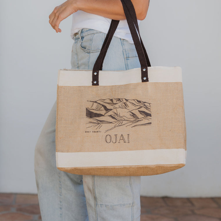Person holding a tote bag with 'Ojai' branding against a neutral background