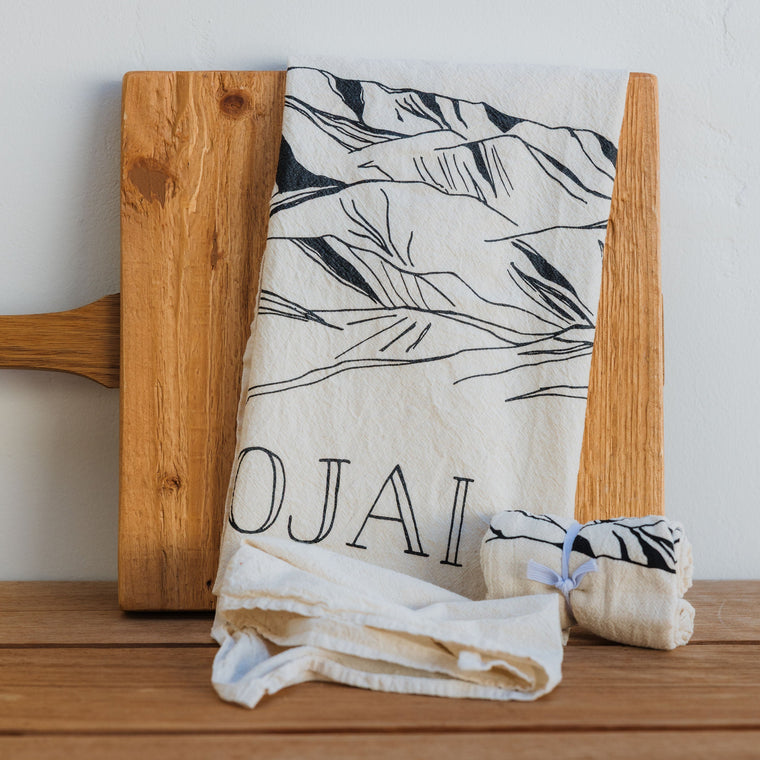 Wooden cutting board with a folded towel featuring 'Ojai' on a wooden surface.