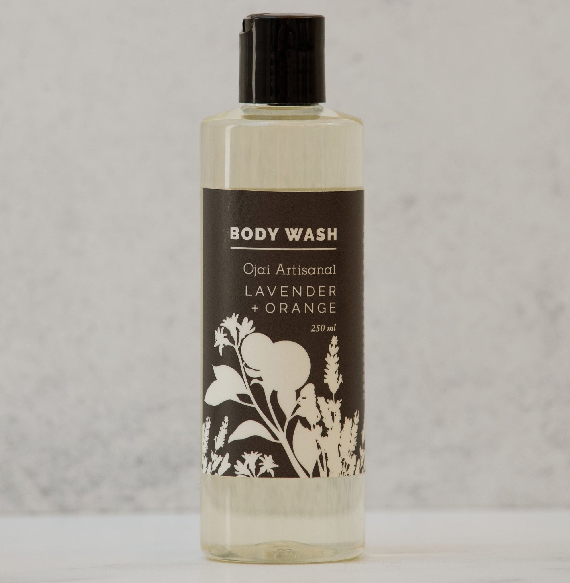 A clear bottle of body wash with a label featuring black and white botanical illustrations, labeled 'Ojai Artisanal Lavender + Orange'.