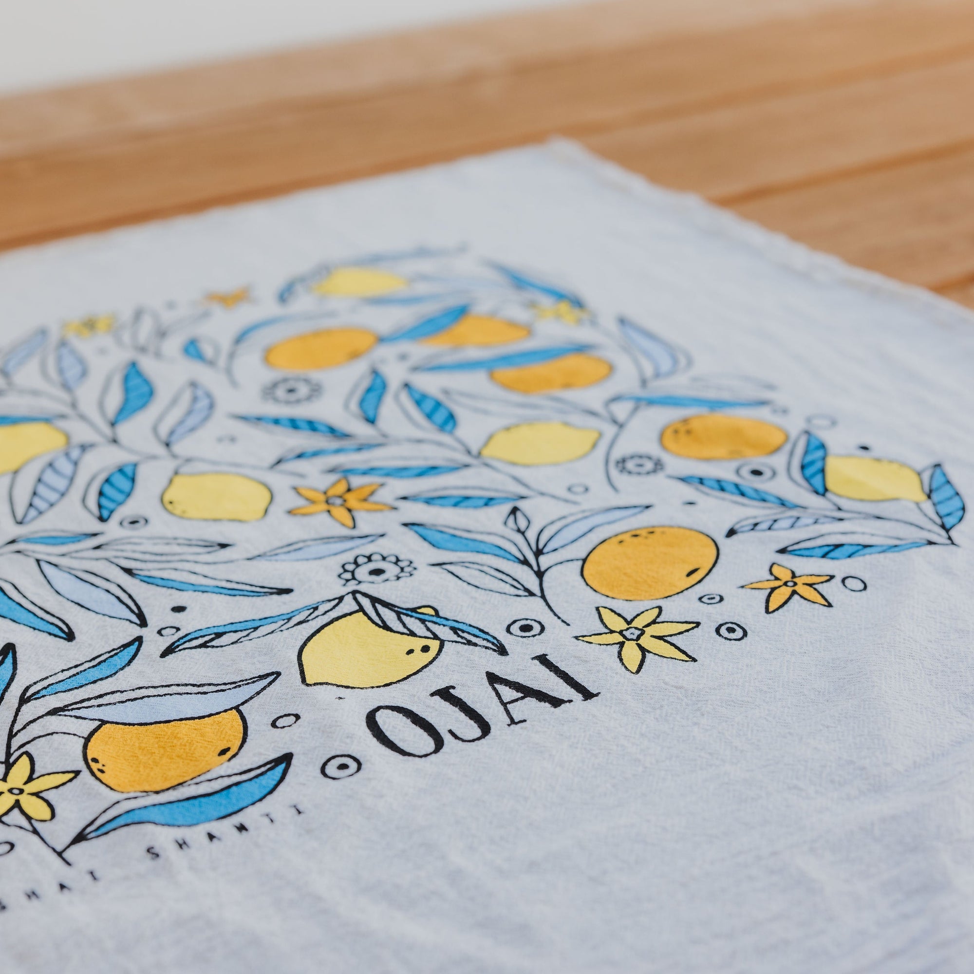 Hand-drawn design of lemons and flowers on a white towel with 'Ojai' printed on it, placed on a wooden surface.
