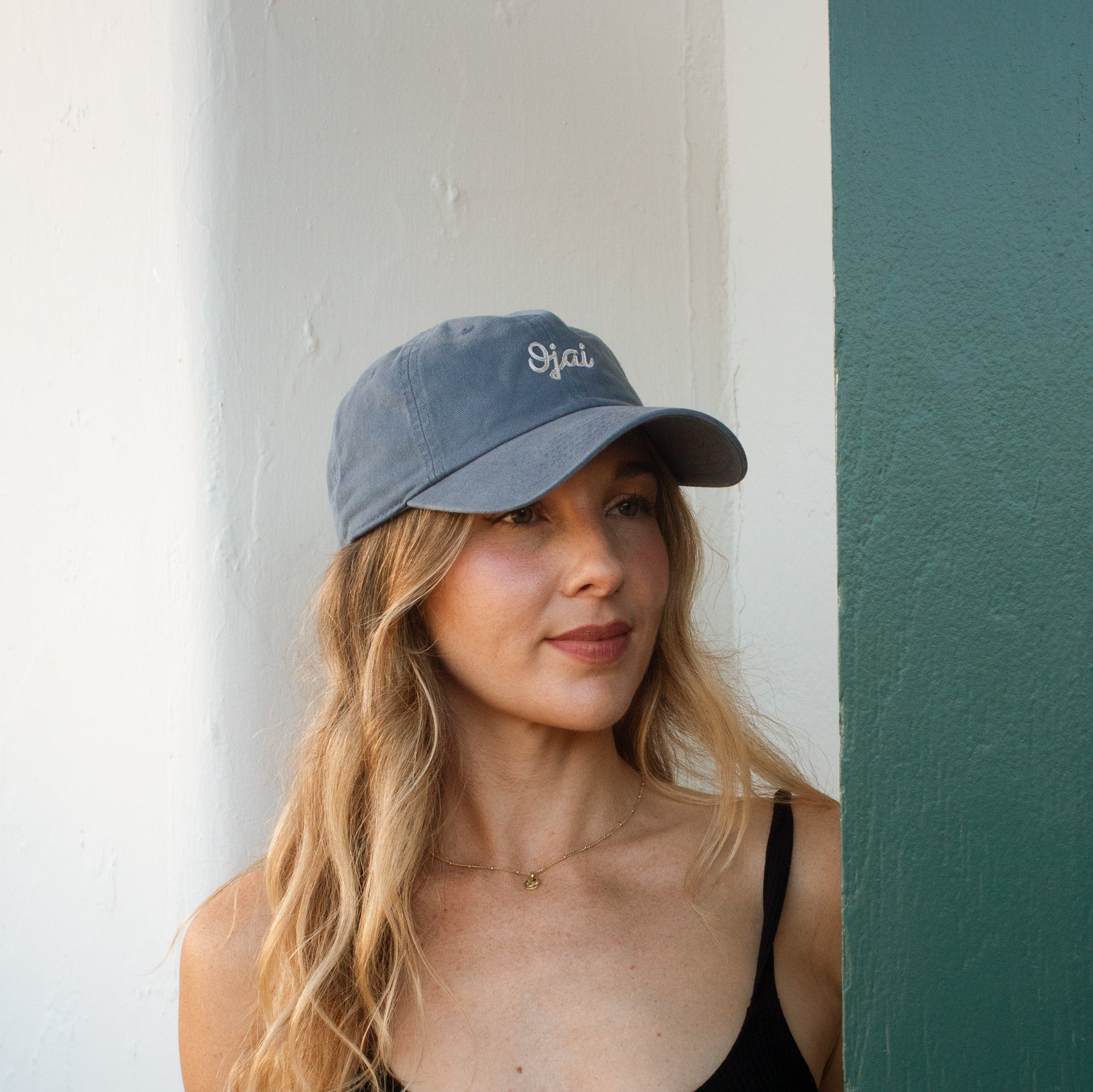 A woman with long blonde hair wearing a slate blue baseball cap with 'Ojai' embroidered in white on the front, leaning against a white wall next to a dark green painted column.