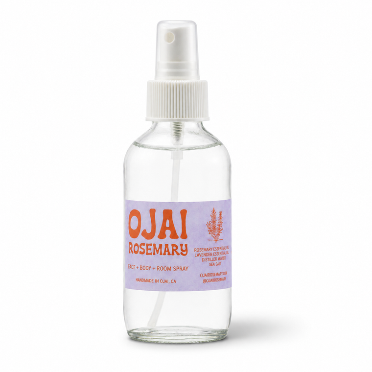 A clear glass spray bottle of Ojai Rosemary room spray with a white pump dispenser and a lavender label featuring orange text, set against a plain white background.