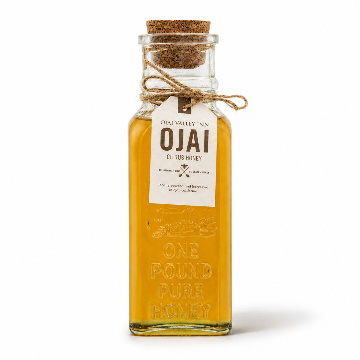 A clear glass bottle filled with golden amber honey, sealed with a cork stopper and tied with twine. A white paper label reads 'OJAI CITRUS HONEY' and embossed on the bottle is 'ONE POUND PURE HONEY.