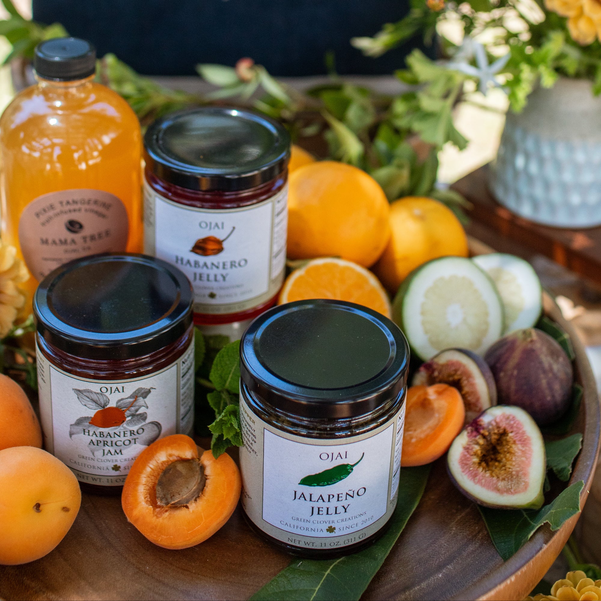 Three jars of OJAI jelly arranged on a wooden board with apricots, figs, and sliced cucumbers, alongside an orange juice bottle and a navy tote bag with OJAI lettering in a garden setting.