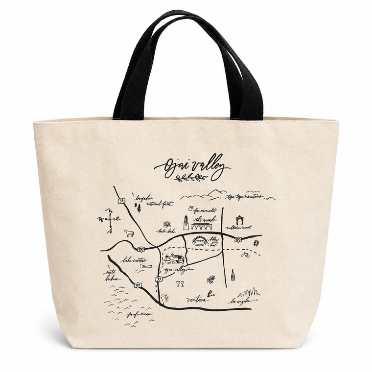 A natural canvas tote bag with black handles, featuring a black hand-drawn map illustration of Ojai Valley with the word 'Ojai Valley' written above it.