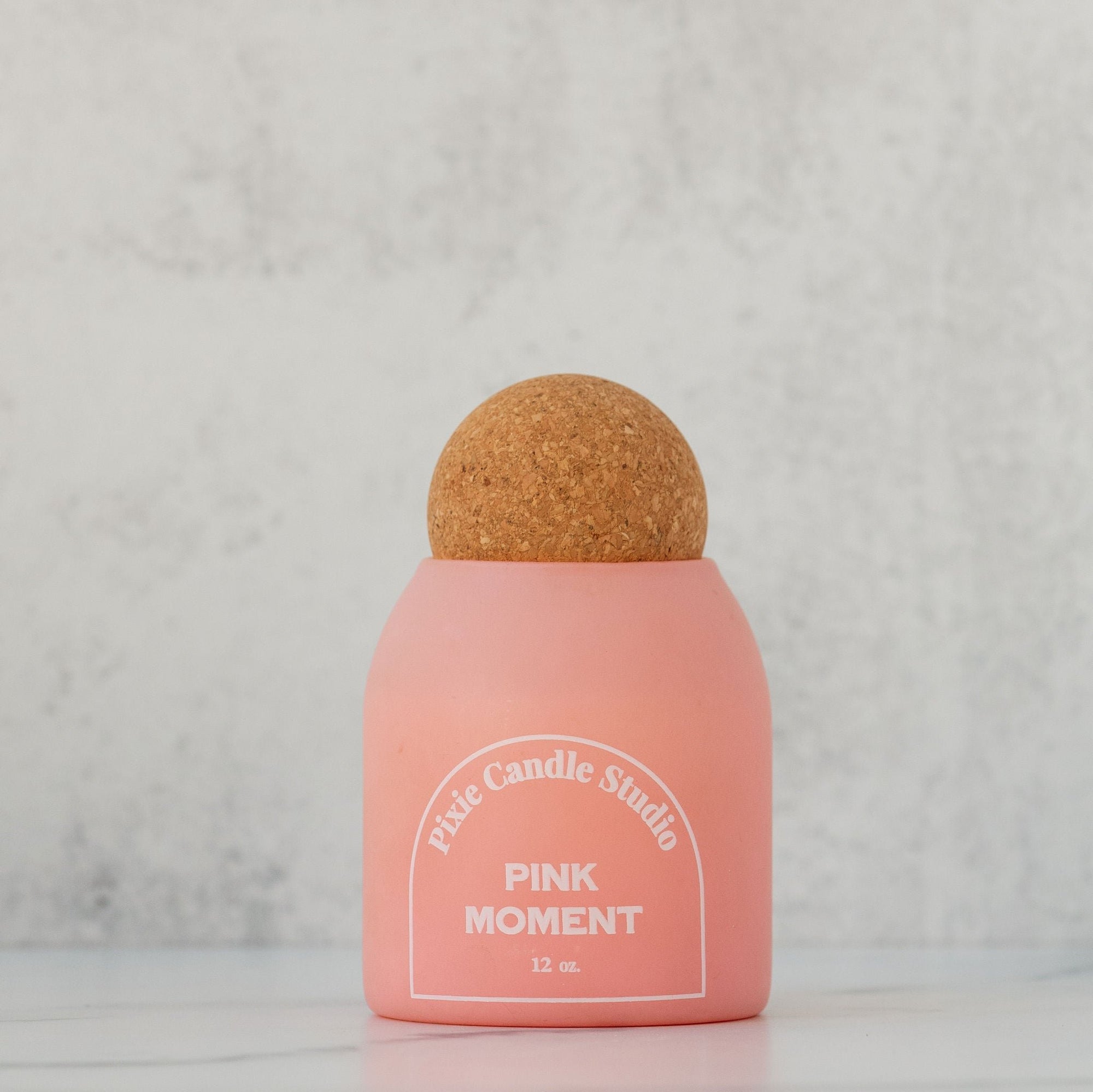 A pink glass jar candle with a brownish-beige wax top, labeled 'Pixie Candle Studio Pink Moment'.