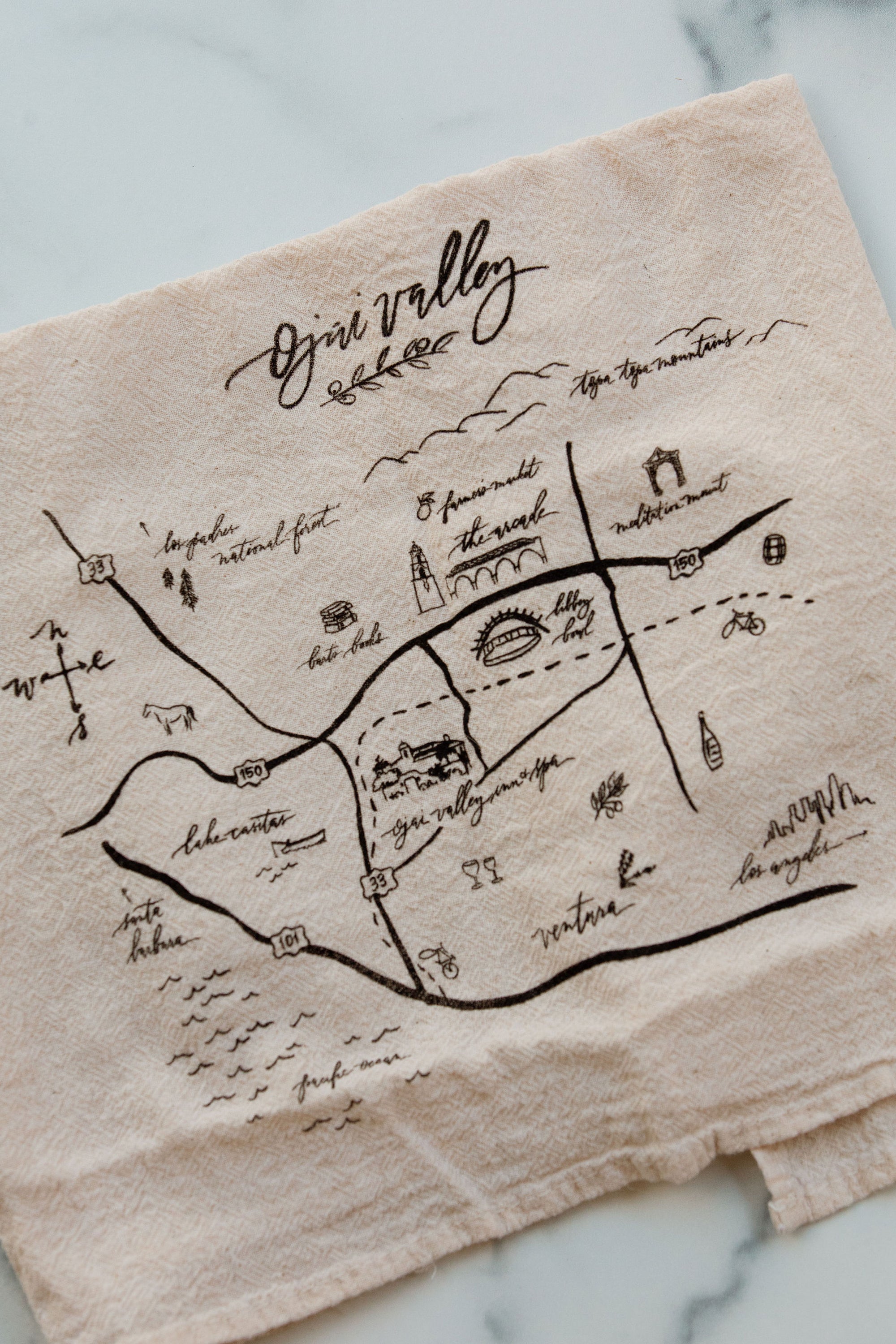 A beige cotton dish towel with a black printed map of the Ojai Valley.