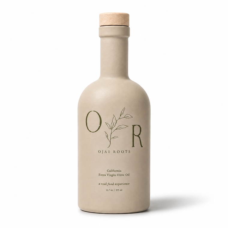 A matte beige ceramic bottle of Ojai Roots Extra Virgin Olive Oil with a natural cork stopper, shown on a clean white background.
