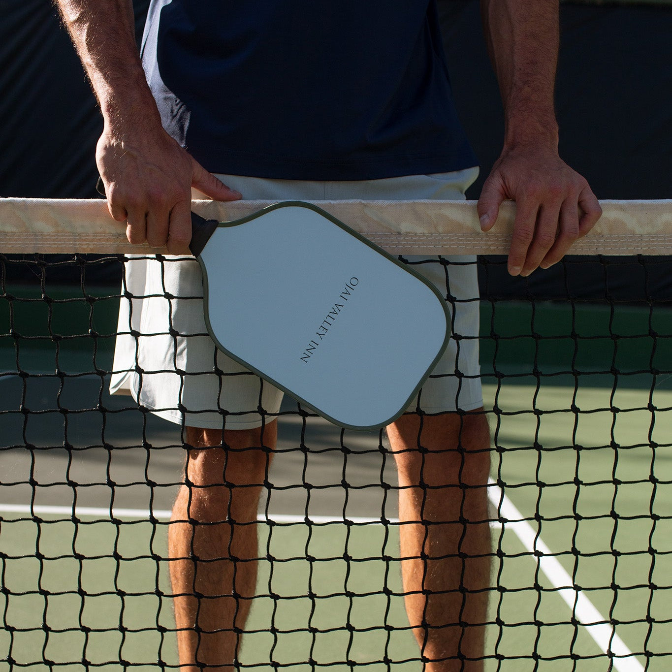 A person standing on a tennis court behind the net, holding a paddle racket. The person is wearing a navy blue shirt, light-colored shorts, and white sneakers.