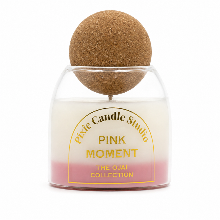 A jar candle with a clear glass body showing a white-to-pink gradient wax, topped with a cork sphere on a metal rod. The front label reads 'Pxic Candle Studio PINK MOMENT THE OJAI COLLECTION'.