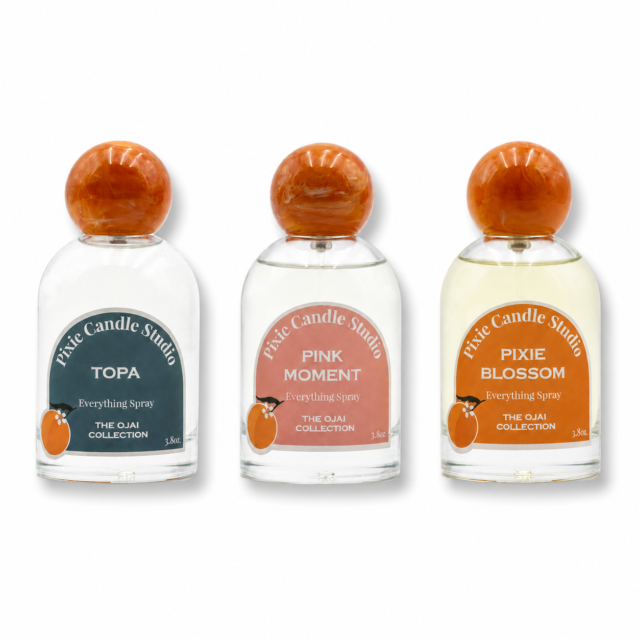 Three small clear glass bottles of Pixie Candle Studio Everything Spray from The Oui Collection, arranged side by side on a white background. The bottles have rounded orange wooden caps and display different colored labels: dark teal for 'Topa', dusty pink for 'Pink Moment', and orange for 'Pixie Blossom'.