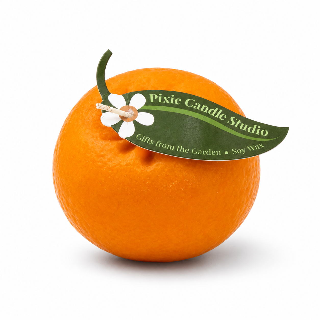 A bright orange citrus fruit with a green stem and a decorative green paper tag reading 'Pixie Candle Studio' attached, photographed on a clean white background.
