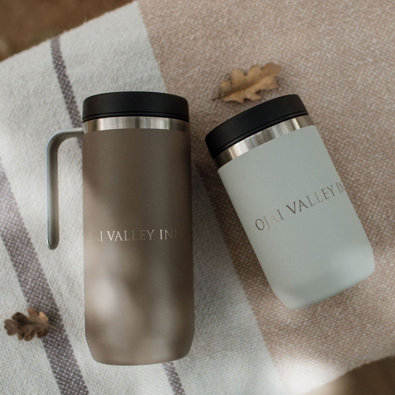 Two insulated travel tumblers with lids from O'Neill Valley Inn are displayed on a striped beige and gray blanket with scattered autumn leaves. One tumbler is matte taupe-brown with a handle, and the other is matte sage green, both featuring silver metallic bands and the brand name 'O'NEILL VALLEY INN' printed on the front.