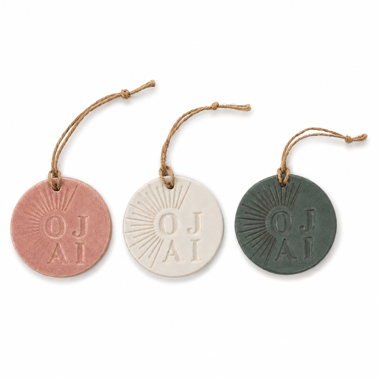 Three round hanging tags or ornaments in dusty pink, cream, and dark green, each embossed with a sunburst and the letters 'OJ AI' and suspended from a short piece of twine, shown on a white background.