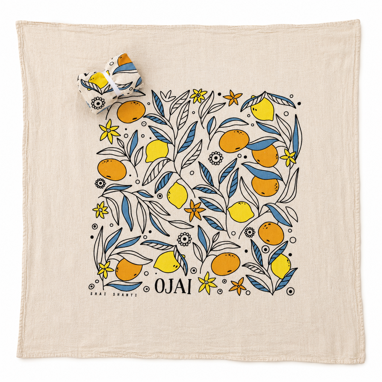 A square cream-colored cotton towel featuring a colorful botanical illustration of oranges, lemons, and blue leaves, with the word 'OJAI' printed at the bottom. A matching rolled-up corner of the towel is shown in the upper left, set against a clean white background.