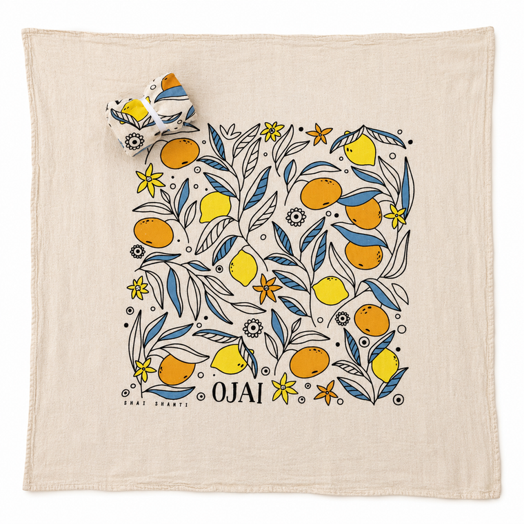 A square cream-colored cotton towel featuring a colorful botanical illustration of oranges, lemons, and blue leaves, with the word 'OJAI' printed at the bottom. A matching rolled-up corner of the towel is shown in the upper left, set against a clean white background.