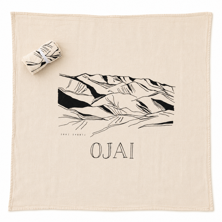 A square cream-colored cotton towel featuring a black line art illustration of desert canyon mountains and the word 'OJAI' printed below. A matching rolled towel with a black-and-white leaf pattern is placed in the upper left corner on a clean white background.