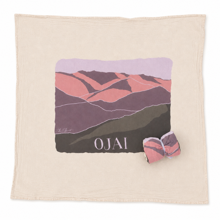 A square cream-colored cotton blanket with a central graphic print of stylized mountains in pink, purple, and green, featuring the text 'OJAI' at the bottom. A matching rolled-up version of the blanket is placed in the lower right corner.