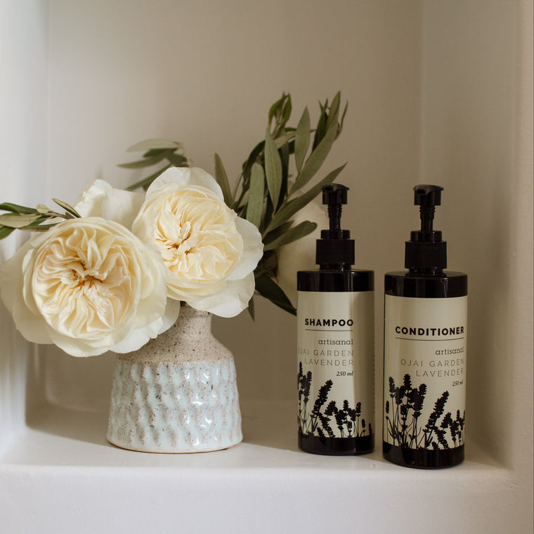 Two pump bottles of shampoo and conditioner with black dispensers and cream labels sit on a white shelf beside a ceramic vase with white flowers and greenery.