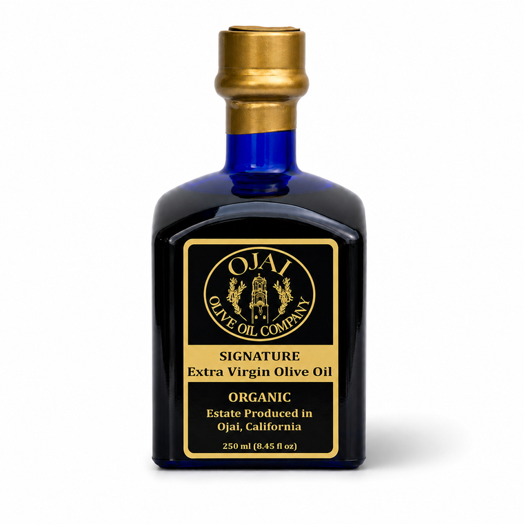 A 250 ml cobalt blue glass bottle of Ojai Signature Organic Extra Virgin Olive Oil, featuring a black label with gold lettering and a gold screw cap, set against a plain white background.