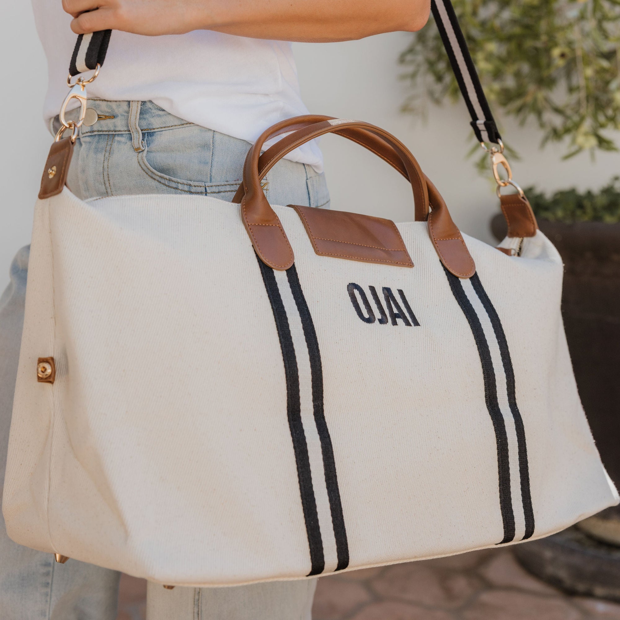 Person holding a beige tote bag with 'Ojai' branding outdoors.