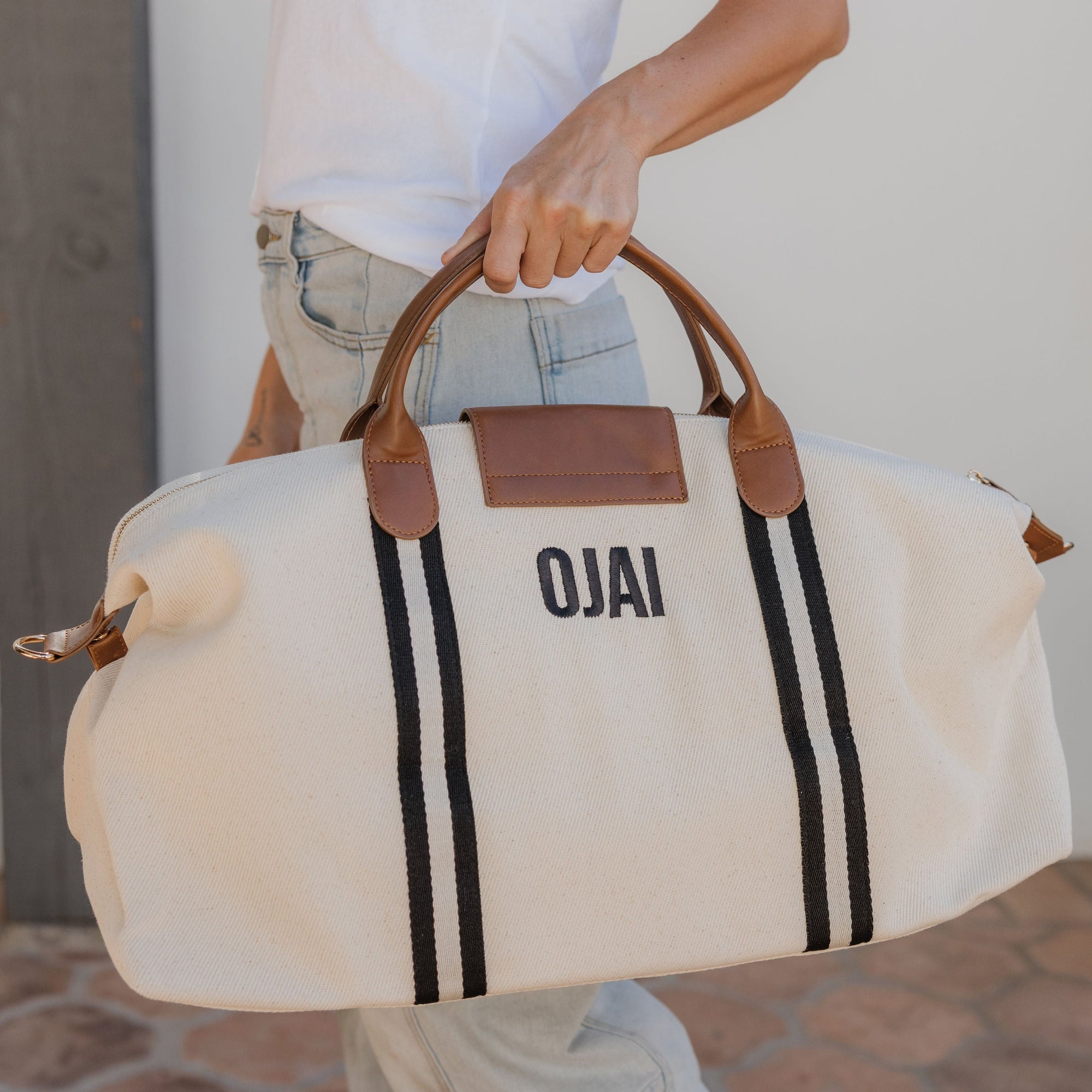 Person holding a beige duffel bag with brown handles and black stripes, featuring the brand 'OJAI'.