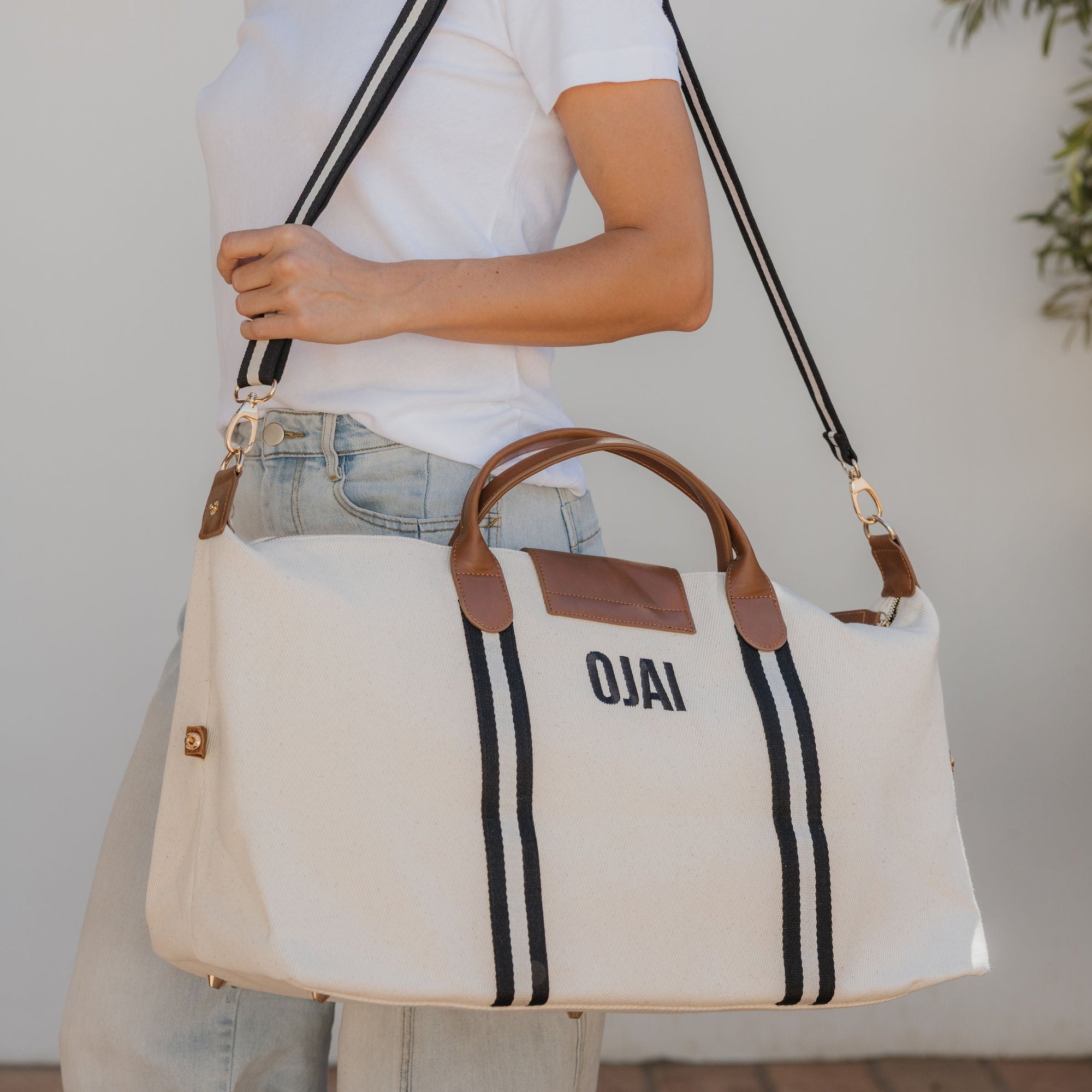 Person holding a white tote bag with brown handles and black stripes, featuring the word 'OJAI'.